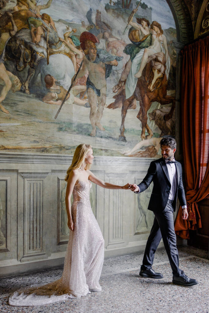Ein formell gekleidetes Paar hält sich an den Händen, während es in einem eleganten Raum neben einem großen historischen Wandgemälde spazieren geht - eine unvergessliche Destination Wedding in Italien.