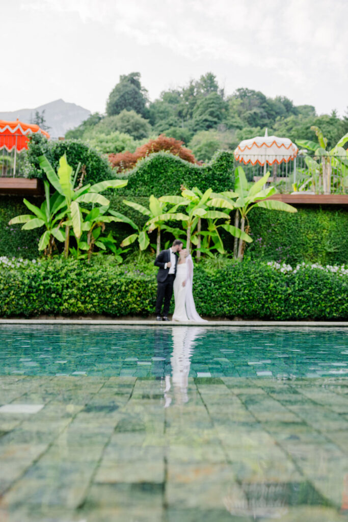Eine Braut und ein Bräutigam stehen bei ihrer verträumten Hochzeit am Pool, umgeben von üppigem Grün und bunten Sonnenschirmen, und fangen die Romantik Apuliens ein.
