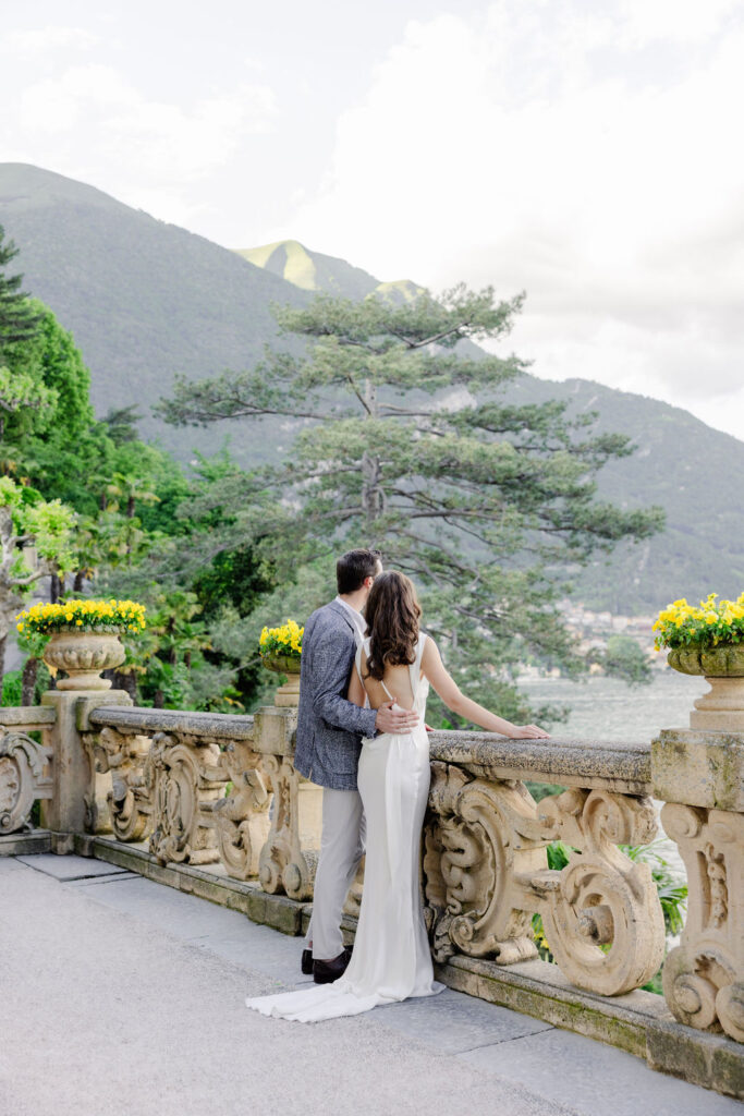 Ein formell gekleidetes Paar steht auf einer Steinterrasse in Italien, mit Blick auf einen See und grüne Berge - eine Traumhochzeit, die von einem erfahrenen Hochzeitsplaner zum Leben erweckt wird.