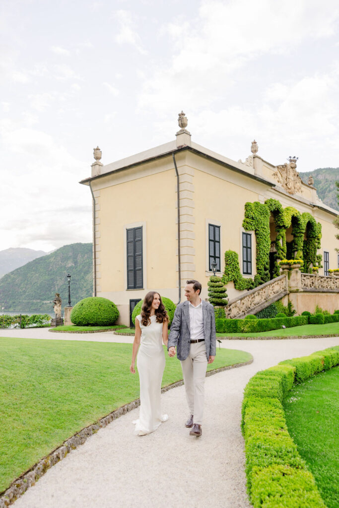 Ein elegant gekleidetes Paar spaziert über einen Gartenweg an einer prächtigen Villa mit gepflegten Hecken und Blick auf die Berge vorbei - eine ideale Kulisse für eine Hochzeit in Italien, die von einem erfahrenen Hochzeitsplaner geplant wurde.