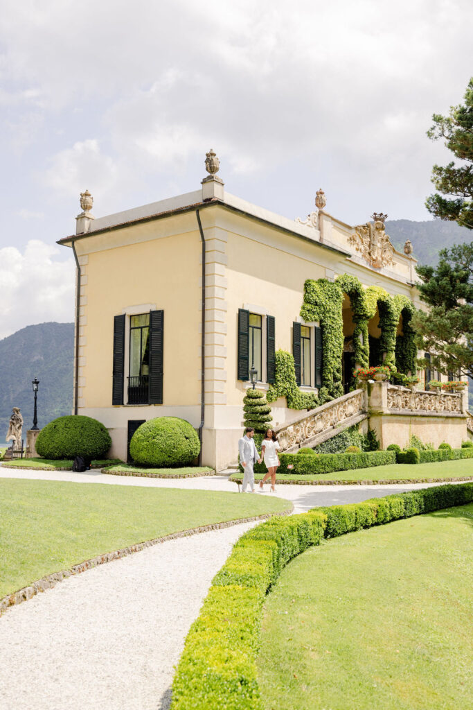 Zwei Personen schlendern unter der italienischen Sonne an einer gelben Villa mit Efeu und gepflegten Gärten vorbei und fangen den Charme Apuliens ein.