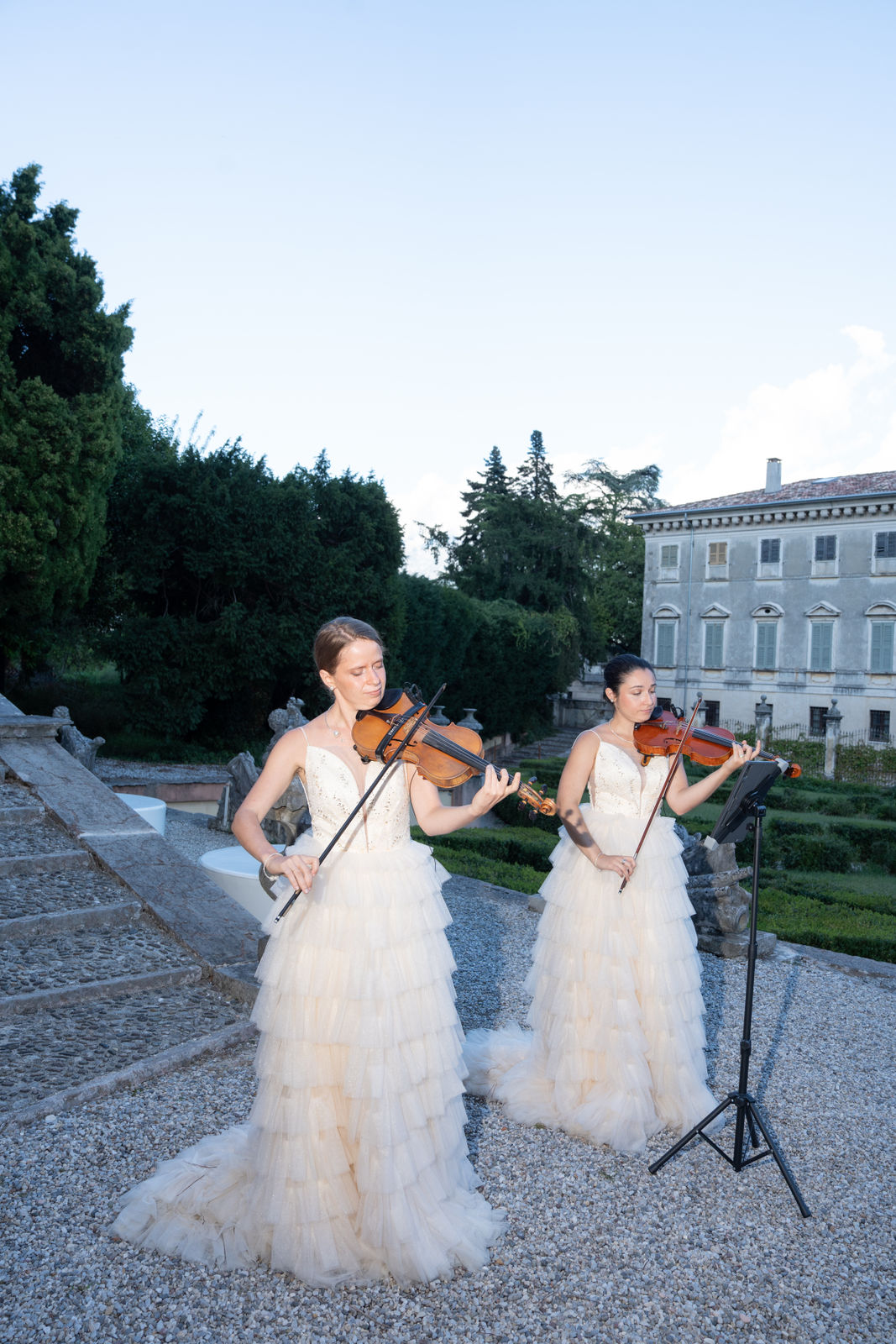 Two violinists in white dresses perform outdoors on Lake Como in front of a stone building.