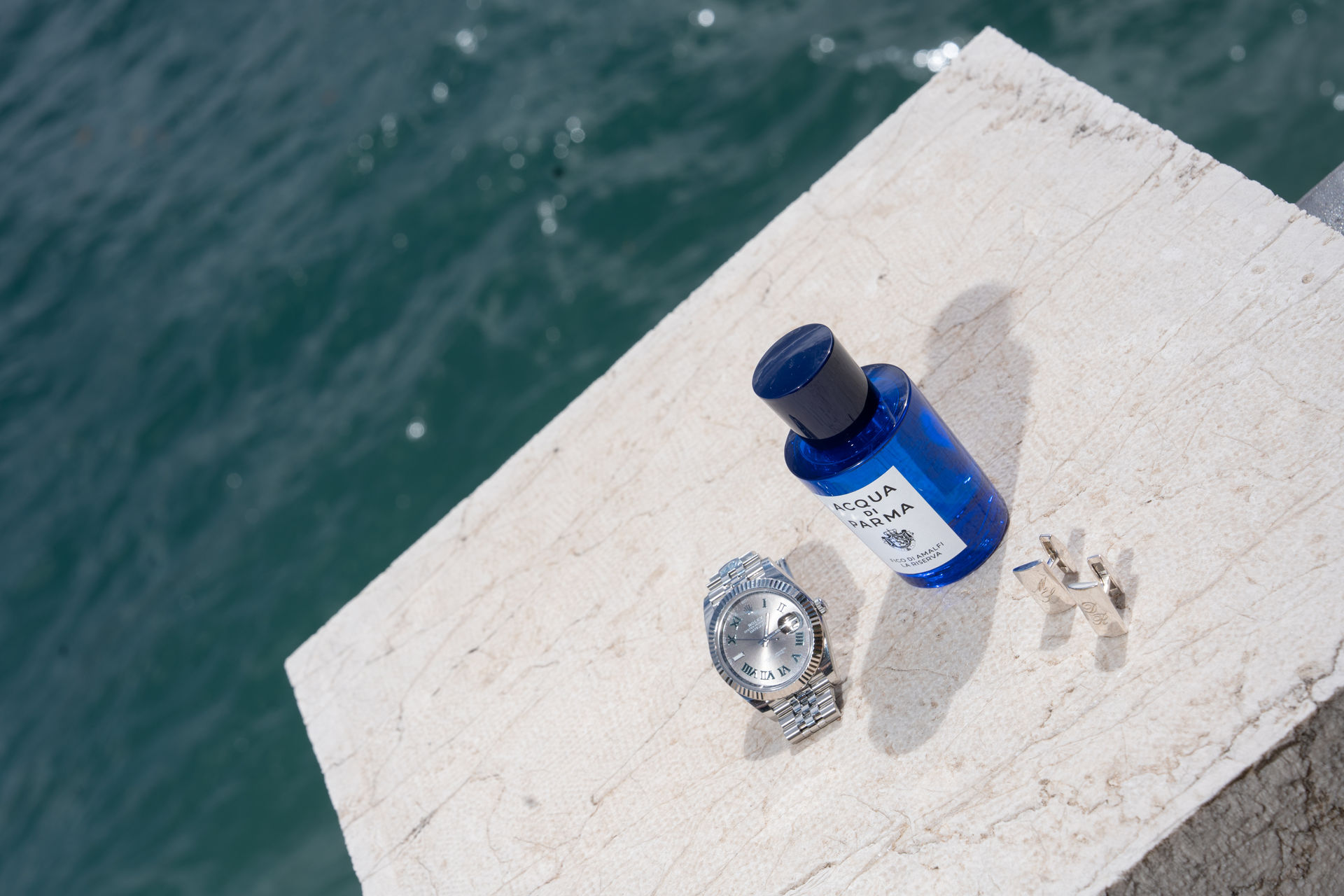 Blaues Parfüm, eine Uhr und Manschettenknöpfe auf einem Felsvorsprung mit Blick auf den Comer See.