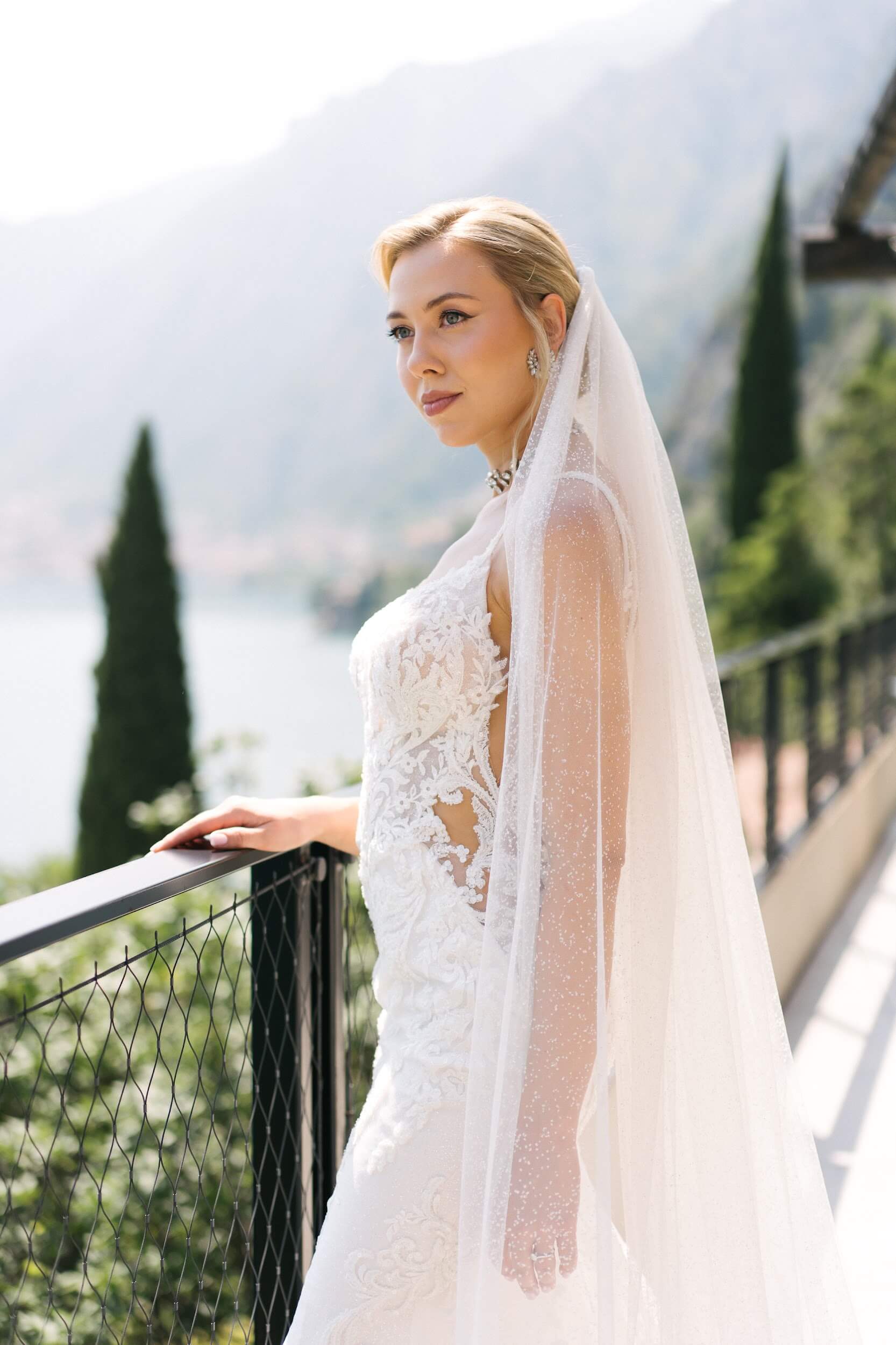 Eine Braut in einem Spitzenkleid und mit Schleier auf einem Balkon in der Toskana mit Blick auf einen See.