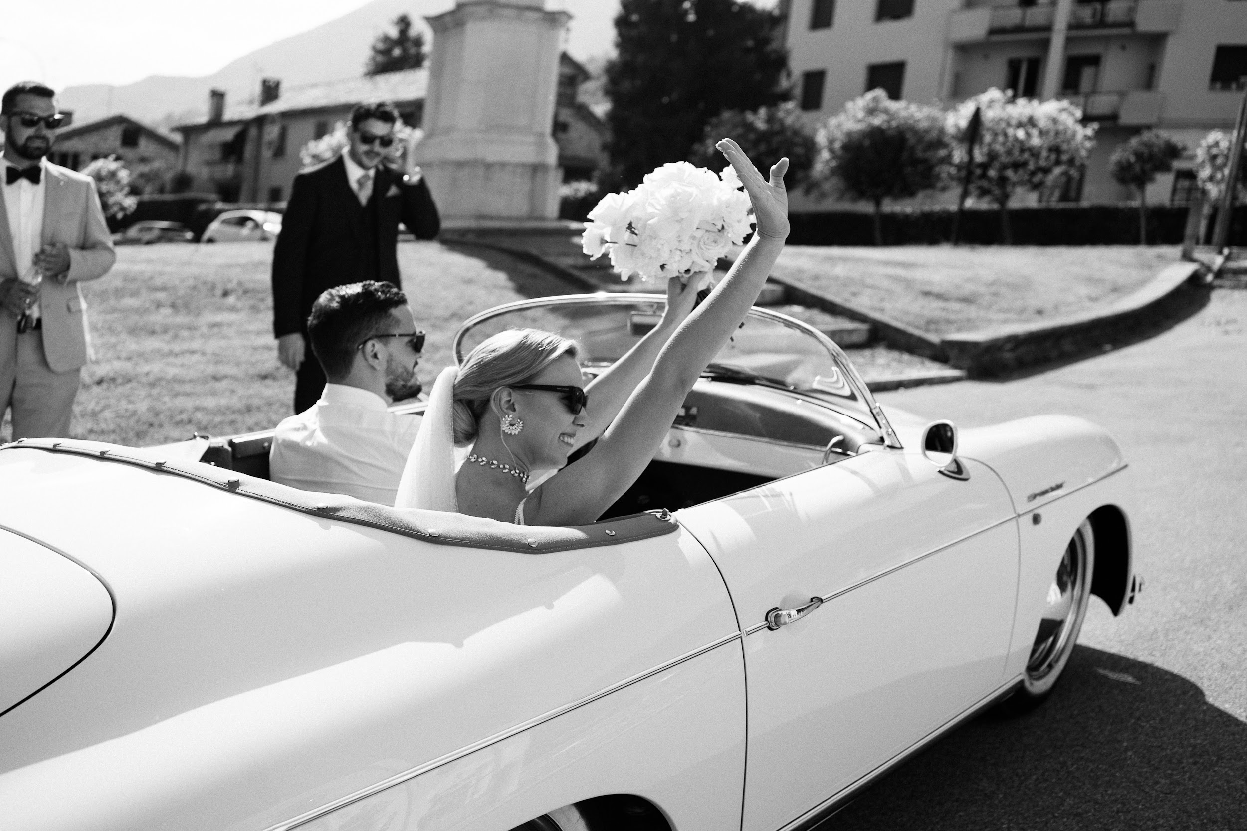 Ein Brautpaar in einem Oldtimer-Cabrio winkt mit seinem Blumenstrauß durch die toskanische Landschaft.