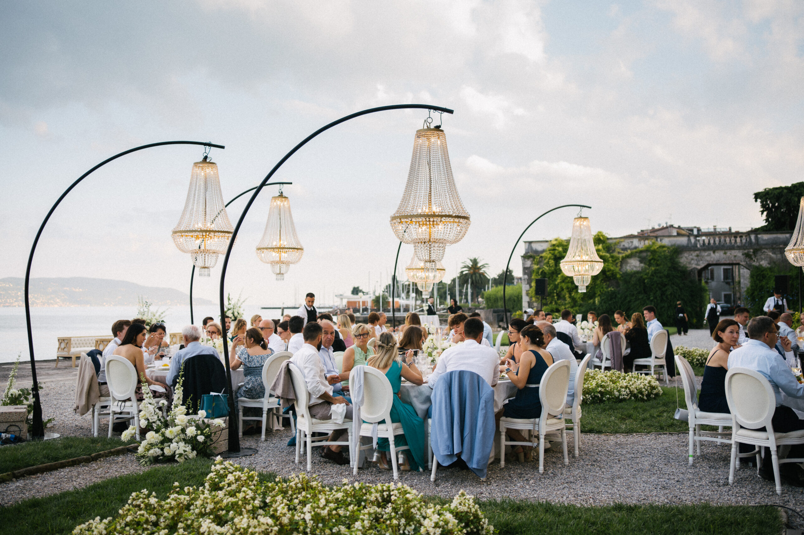 Gäste einer exklusiven Hochzeit speisen im Freien unter Kristallkronleuchtern an einem toskanischen See.