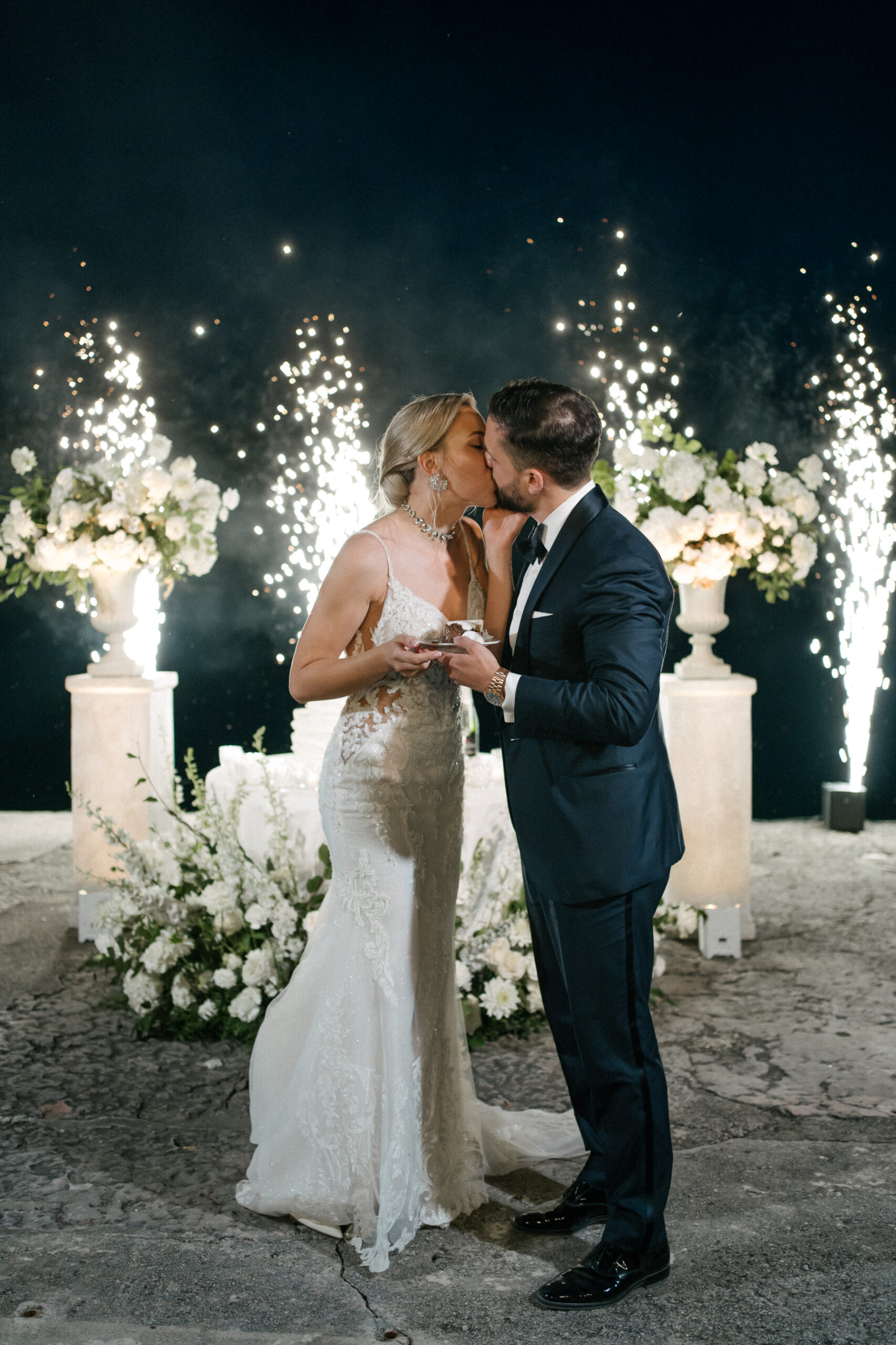 Ein Brautpaar küsst sich bei einer exklusiven Hochzeit in Italien, während es eine Torte in den Händen hält; im Hintergrund ist ein Feuerwerk zu sehen.