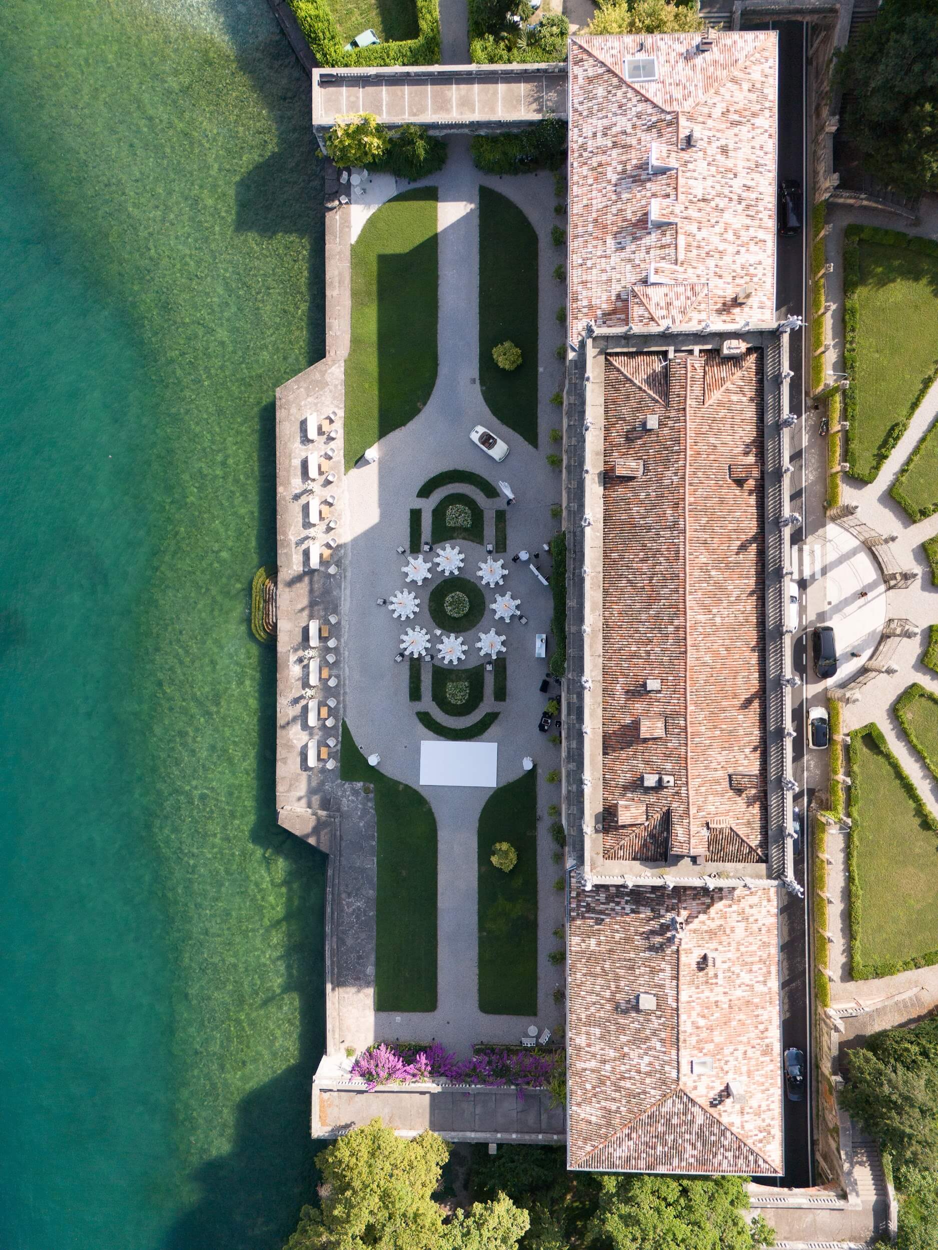 Luftaufnahme eines langgestreckten Gebäudes am See mit einer Gartenterrasse, die für exklusive Hochzeiten hergerichtet ist.